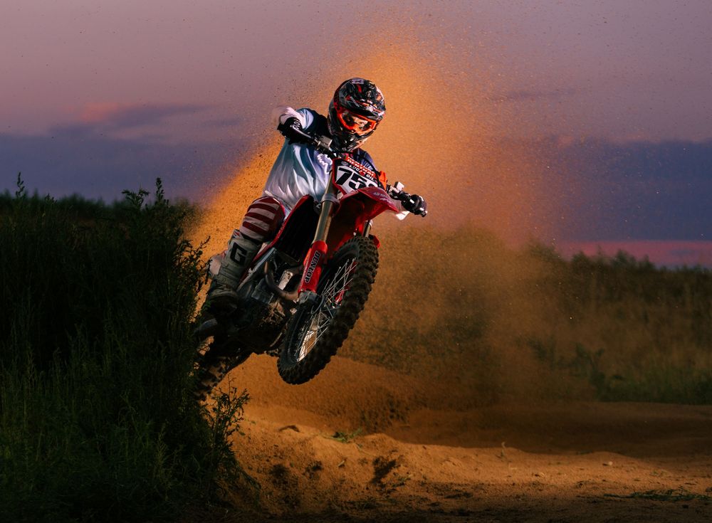 Motocross rider jumps dirt bike against dramatic sunset sky with orange dust cloud trailing behind.