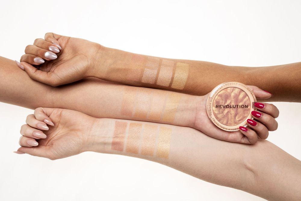 Three diverse skin tones swatched with Revolution highlighter shades, displayed alongside an open compact on white background.