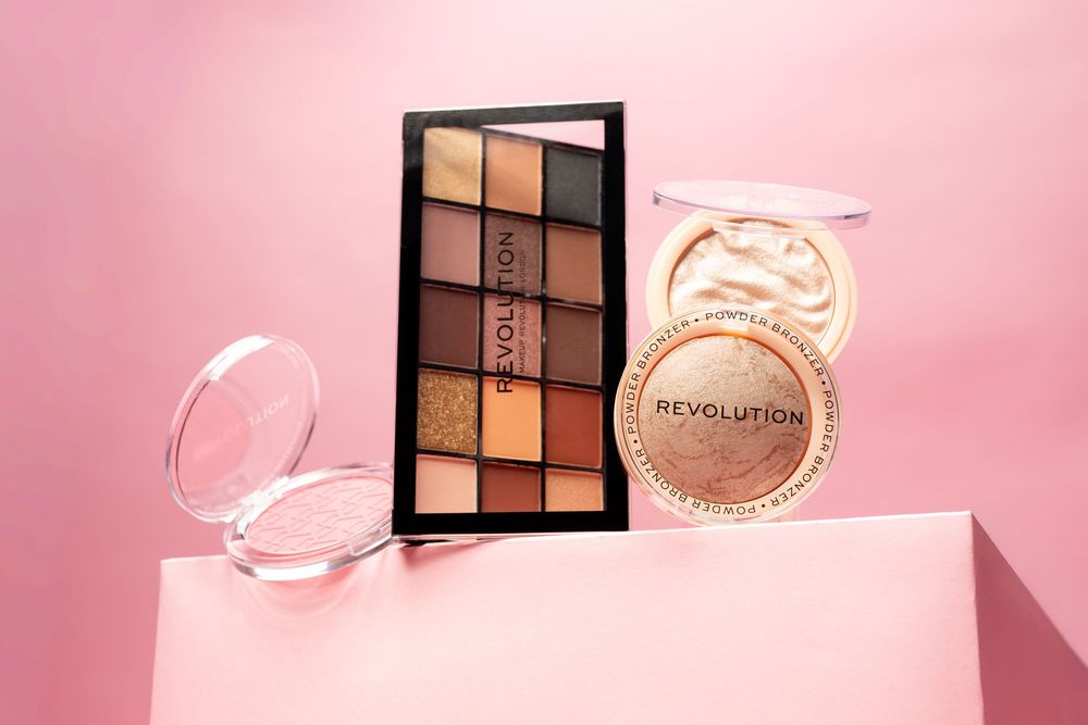 Revolution Beauty makeup palette, highlighter, and compact mirror displayed on pink background.