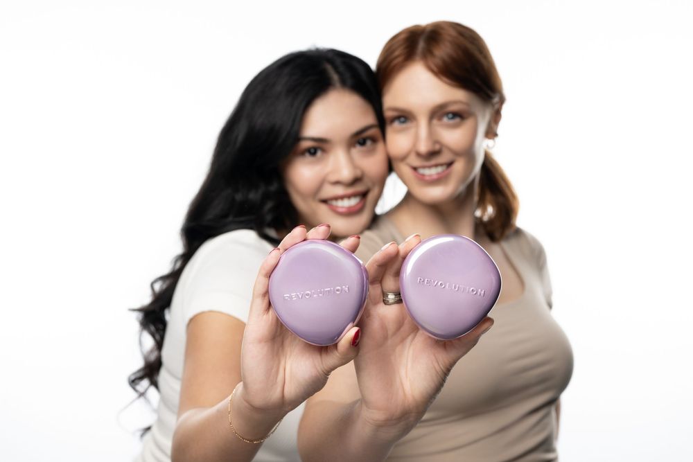 Two women smiling and holding purple compact beauty products against a white background.