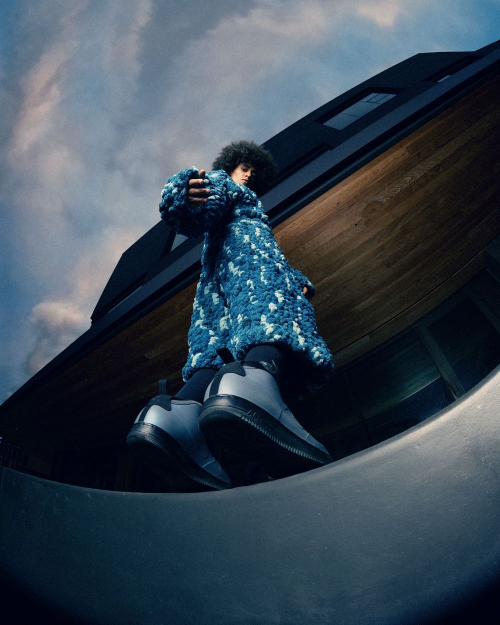 Person in blue floral coat and black boots posing dramatically on curved architecture against cloudy sky.
