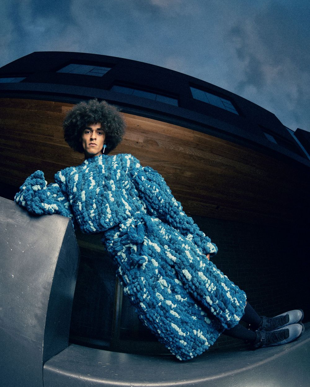 Person wearing a textured blue floral coat with an afro hairstyle, posed dramatically against a curved metallic structure.