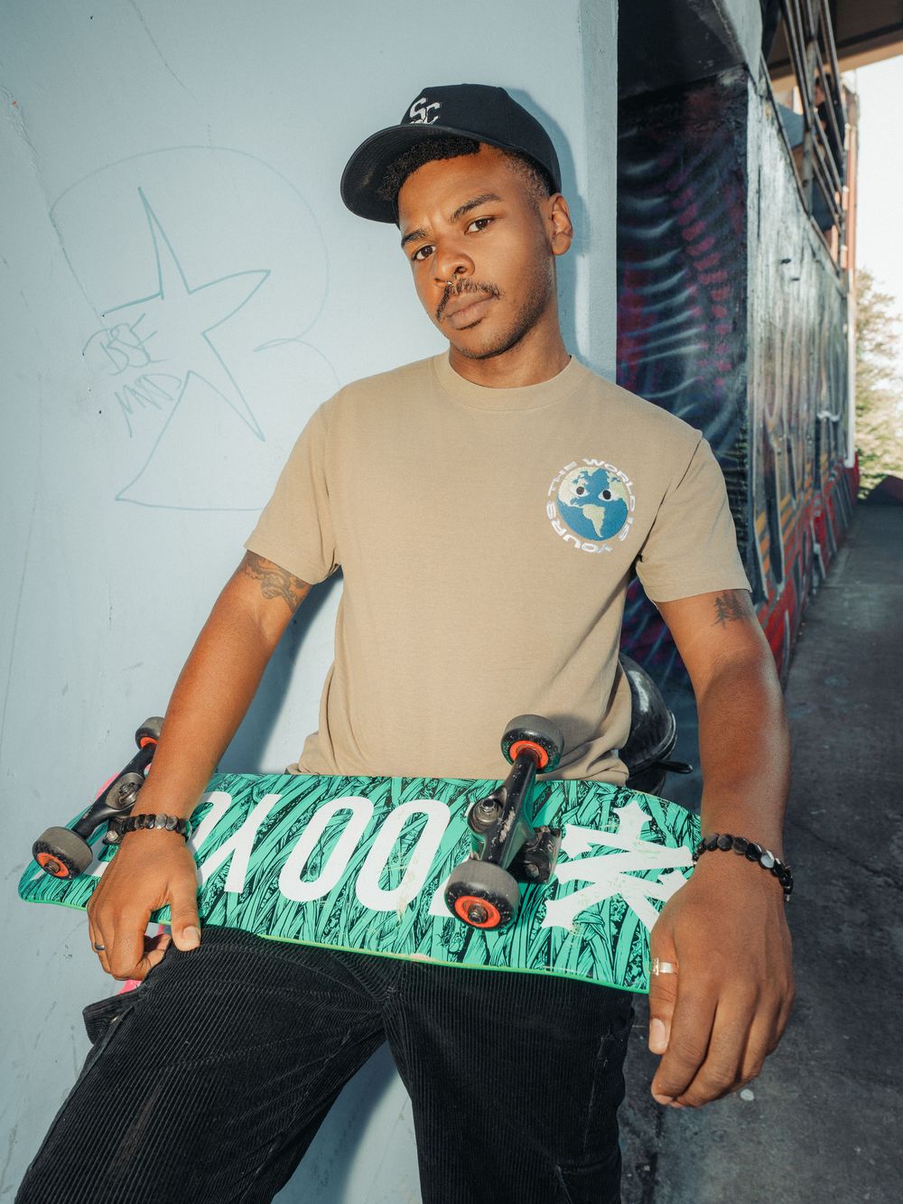 Person wearing a cap and beige t-shirt displays a green skateboard deck with white text graphics in a graffiti setting.