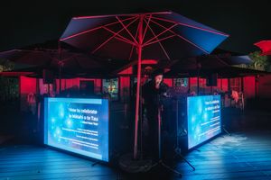 Outdoor rooftop bar with glowing blue LED display panels and red-lit umbrellas at night.