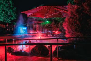 Outdoor nighttime venue with red lighting, mist, large umbrella, rocks, and glowing screen amid lush trees.