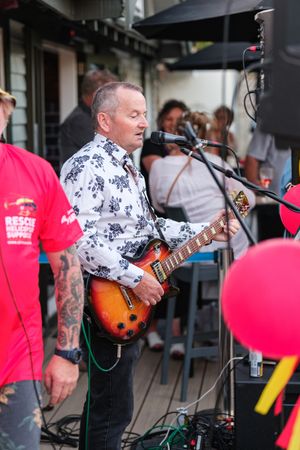Middle-aged guitarist in floral shirt performs live on outdoor stage with electric guitar at a casual music event.