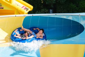 Two people riding a blue tube down a water slide at an outdoor water park on a sunny day.
