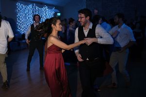 Couple dancing together at a formal event, woman in red dress, man in vest, with fairy lights in background.