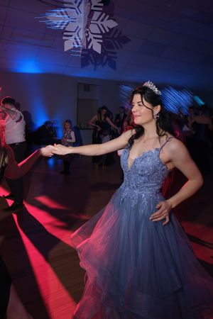 Woman in blue lace ball gown and tiara dancing gracefully under snowflake decorations and colorful lights.