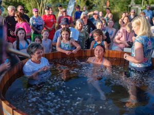 People gathered outdoors watching two women being baptized in a wooden hot tub during a religious ceremony.