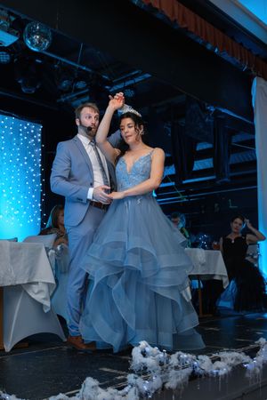 Couple dancing on stage, woman in blue ball gown with tiara, man in gray suit at formal celebration.