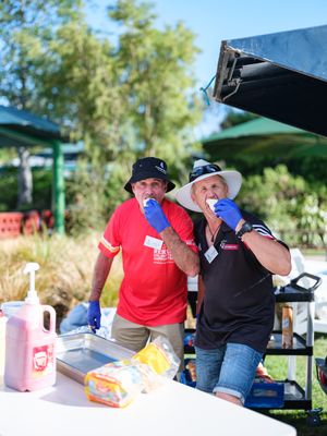 Two volunteers in blue gloves smile while eating at an outdoor community BBQ event with condiments on a white table.