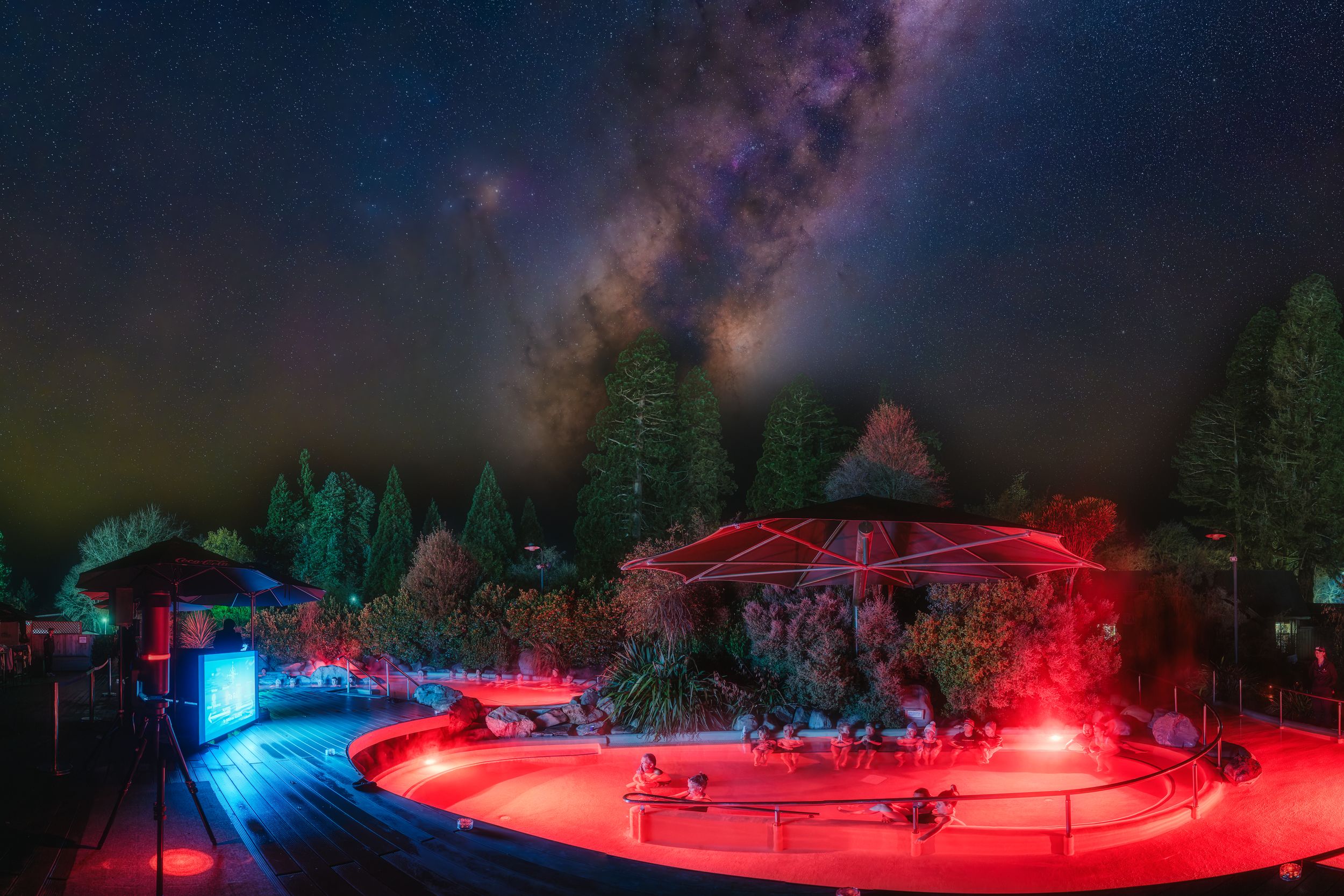 Outdoor amphitheater glowing red under a stunning Milky Way night sky surrounded by tall trees.