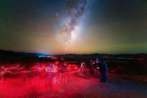 Astronomers observe the Milky Way galaxy with telescopes under a stunning starry night sky illuminated by red lights.