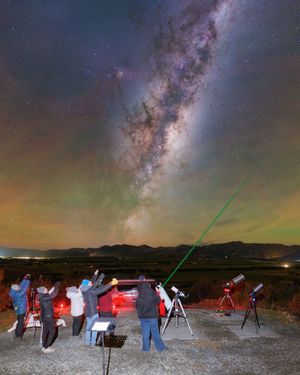 Group of astronomers observing the Milky Way galaxy at night with telescopes and a green laser pointer under a stunning starry sky.