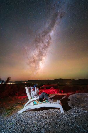 Person reclining on lounge chair under stunning Milky Way galaxy in dark desert sky at night.