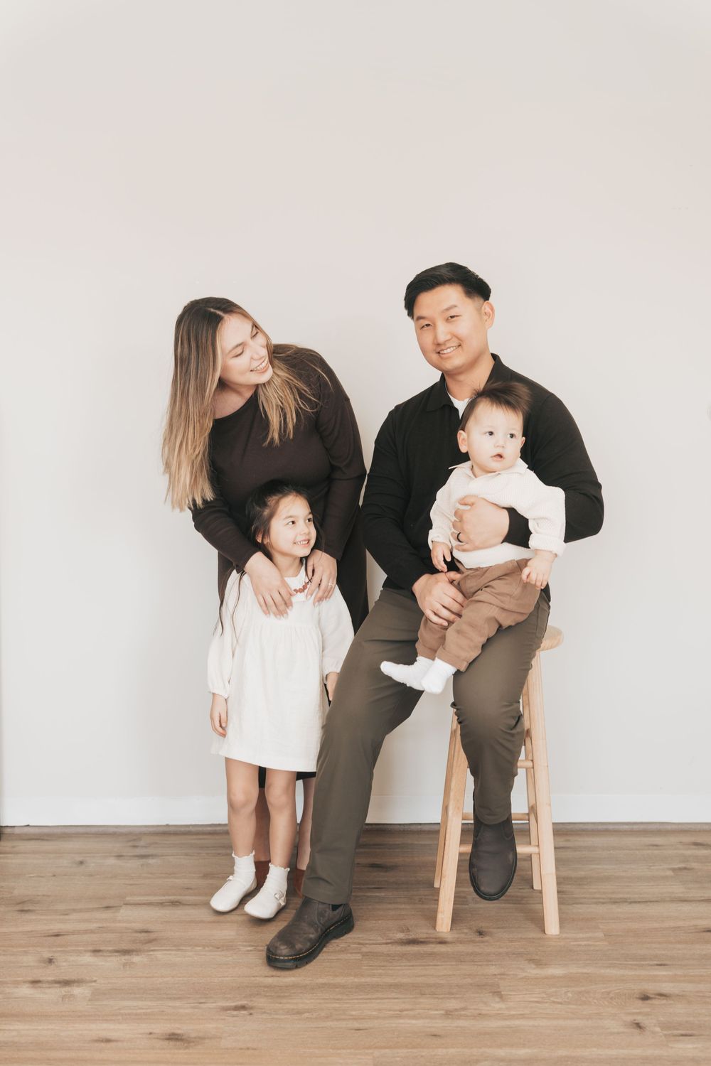 A smiling Asian family of four poses for a portrait, with parents and two young children against a white wall.