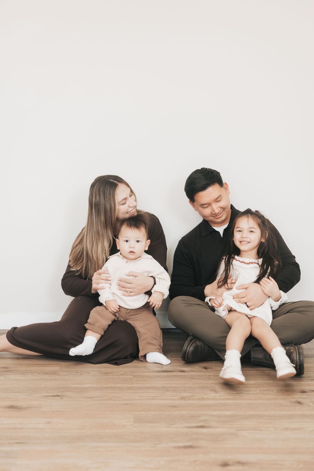 Happy Asian family of four sitting on hardwood floor, parents holding toddler and baby in matching dark outfits.