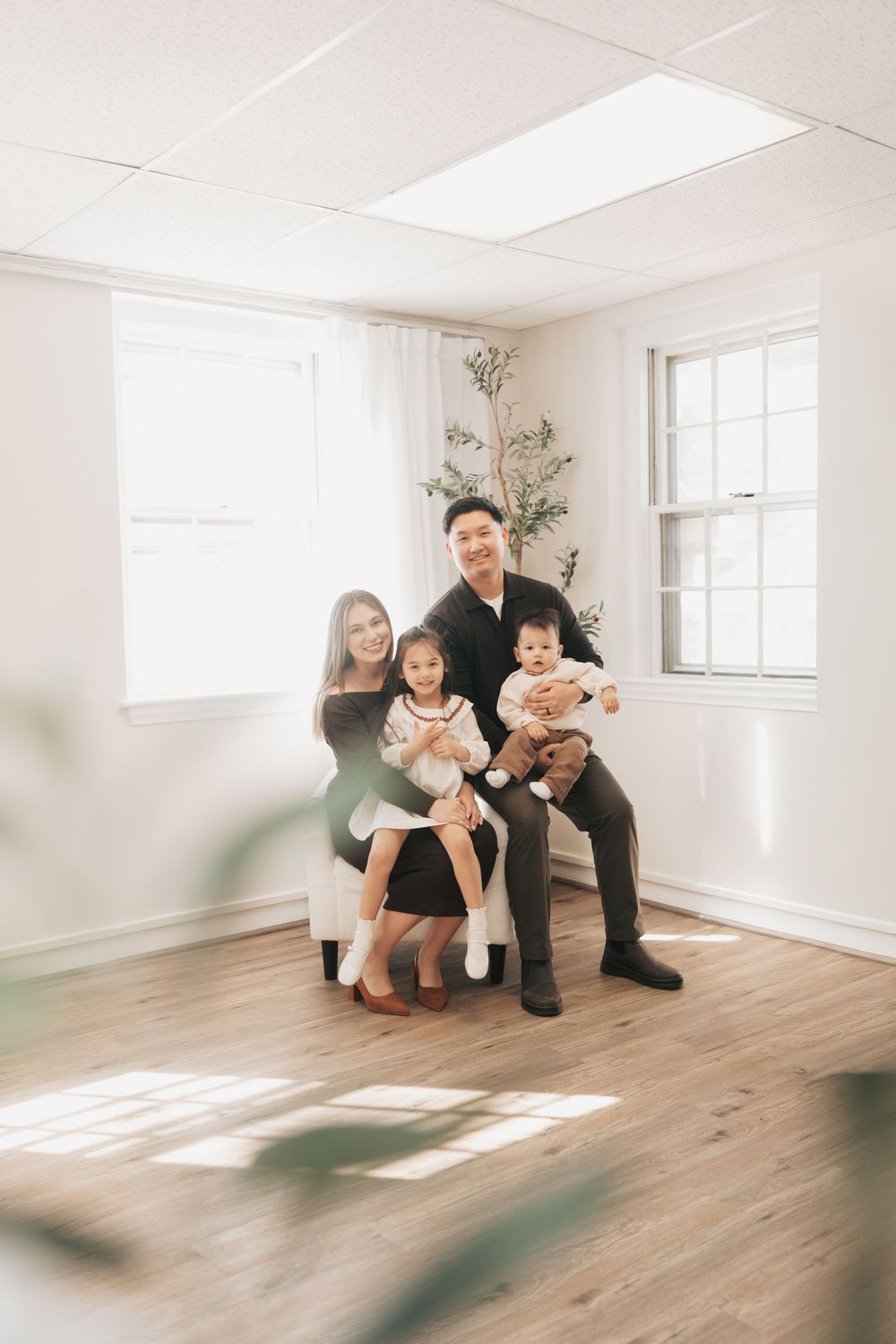 A happy Asian family of four poses together in a bright, minimalist room with hardwood floors and natural light.