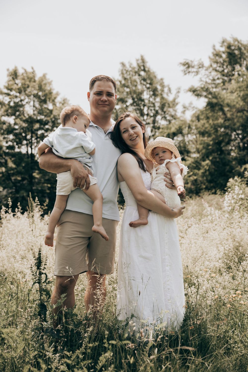 Family wearing white and beige outfits poses together in a sunny meadow with tall grass and trees in the background.