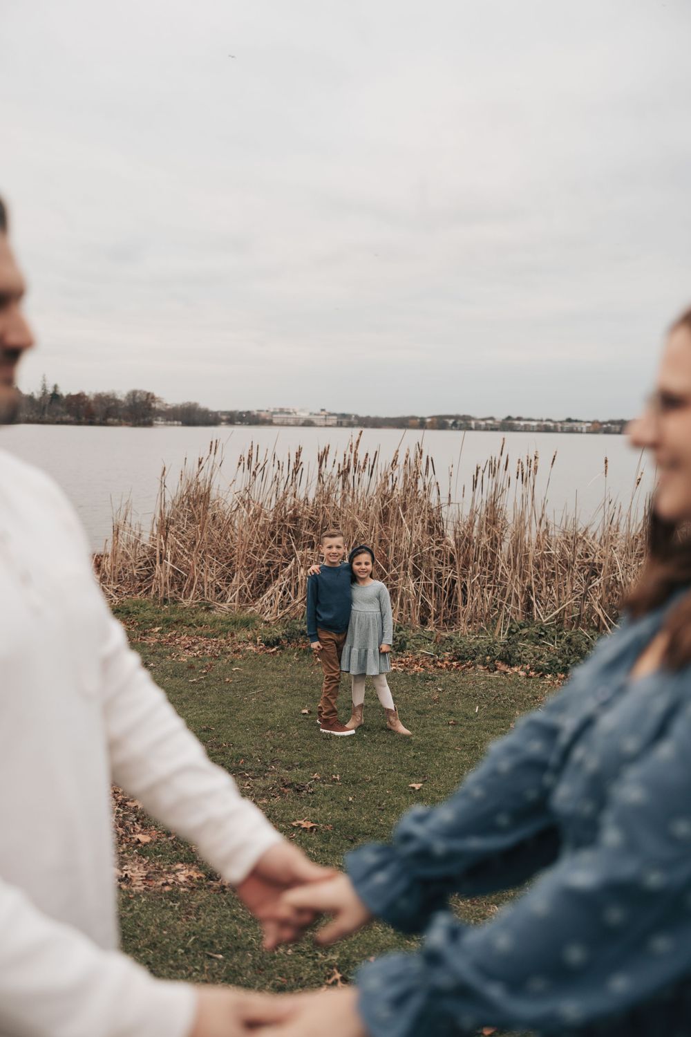 A group holds hands while standing near a lake with cattails and grass on an overcast day.