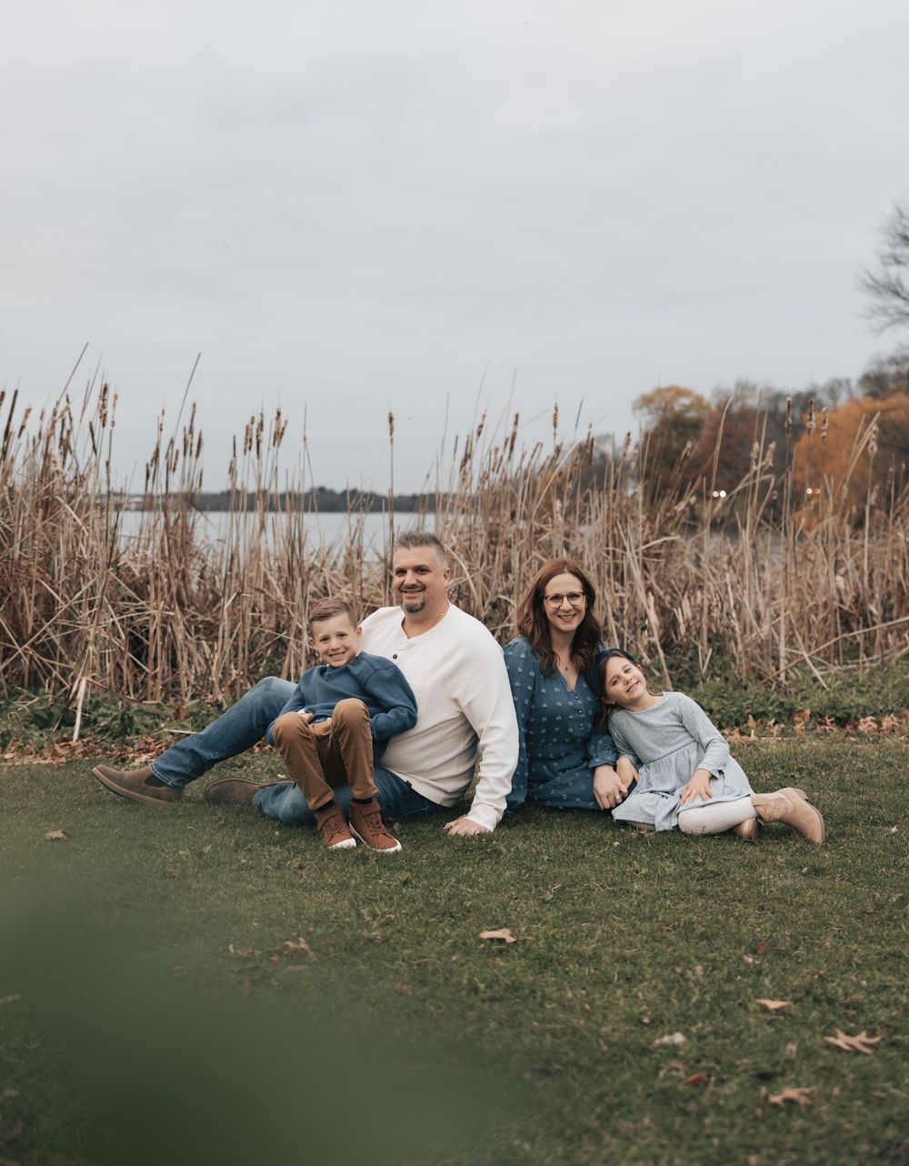 Family relaxing together on grassy hill during autumn afternoon photoshoot with tall dried grasses in background.