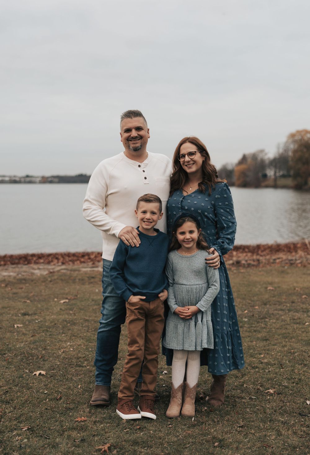 A family of four poses together on an autumn day by a lake, wearing coordinating navy blue and neutral tones.