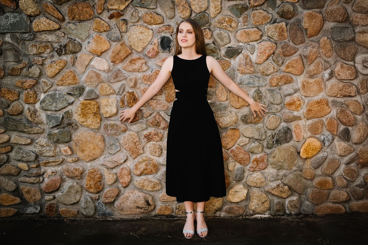 Figure in black dress posed against rustic stone wall background.
