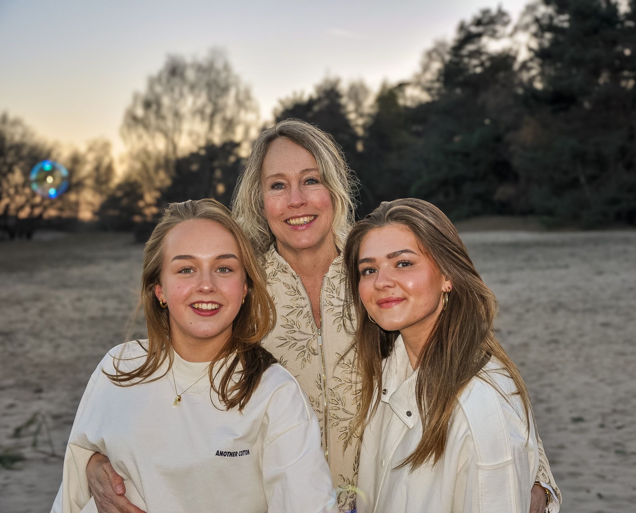 Three smiling women in white sweaters pose together on a sandy beach at sunset.