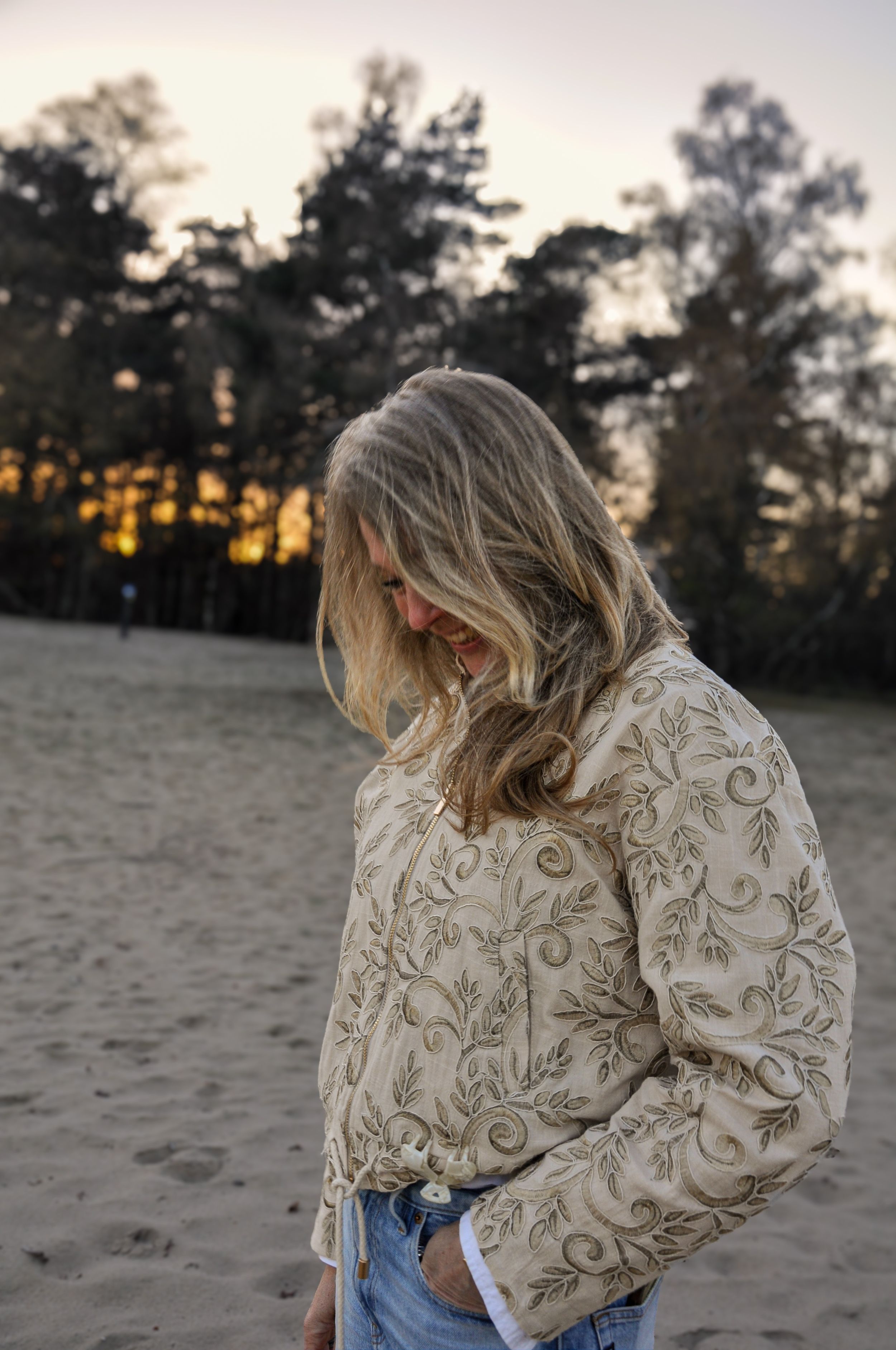 Blonde woman in embroidered cream jacket stands on sandy beach at dusk.