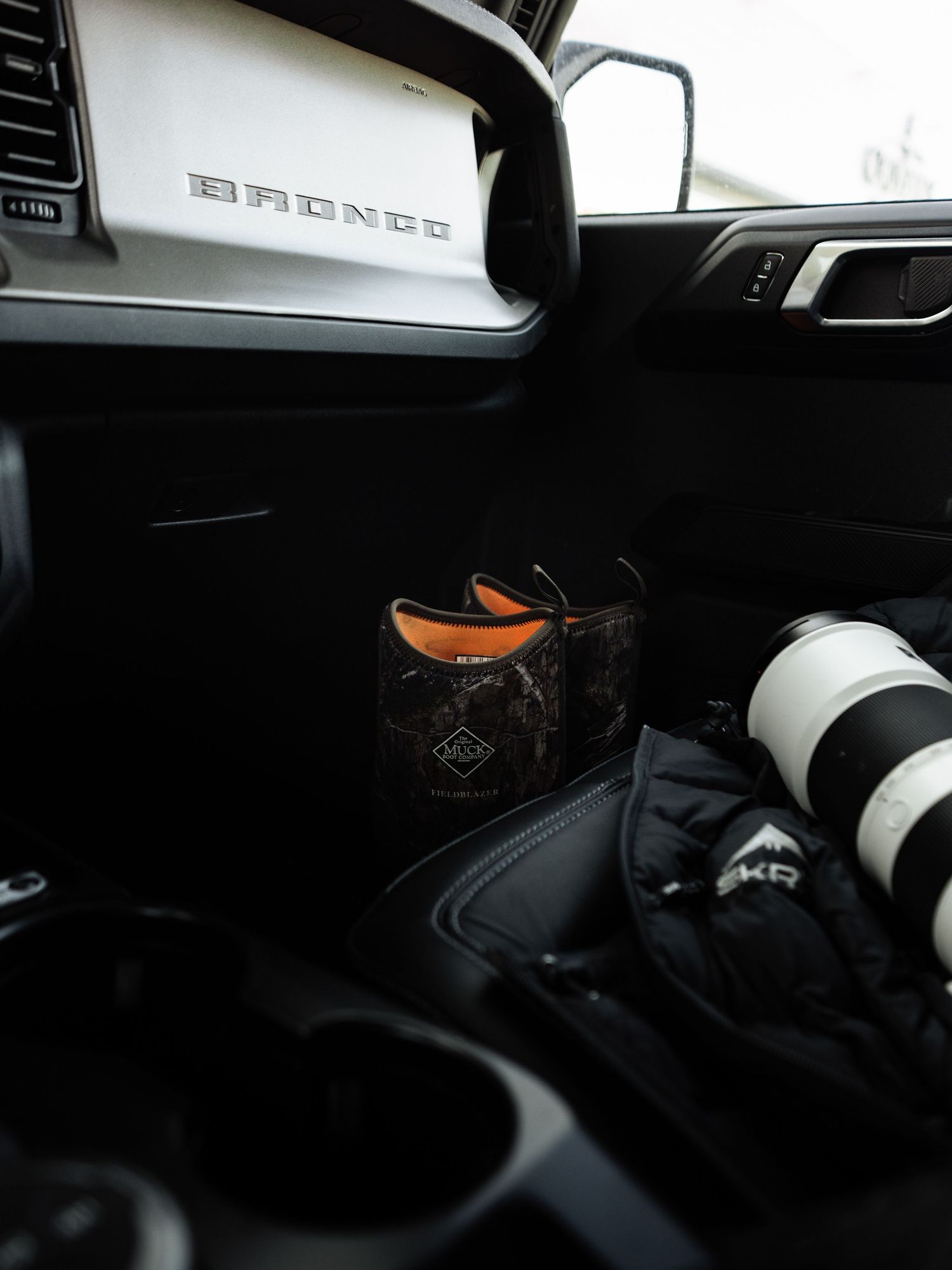Interior of a Ford Bronco with a camo cup holder insert, orange-accented accessory, and camera lens on the center console.