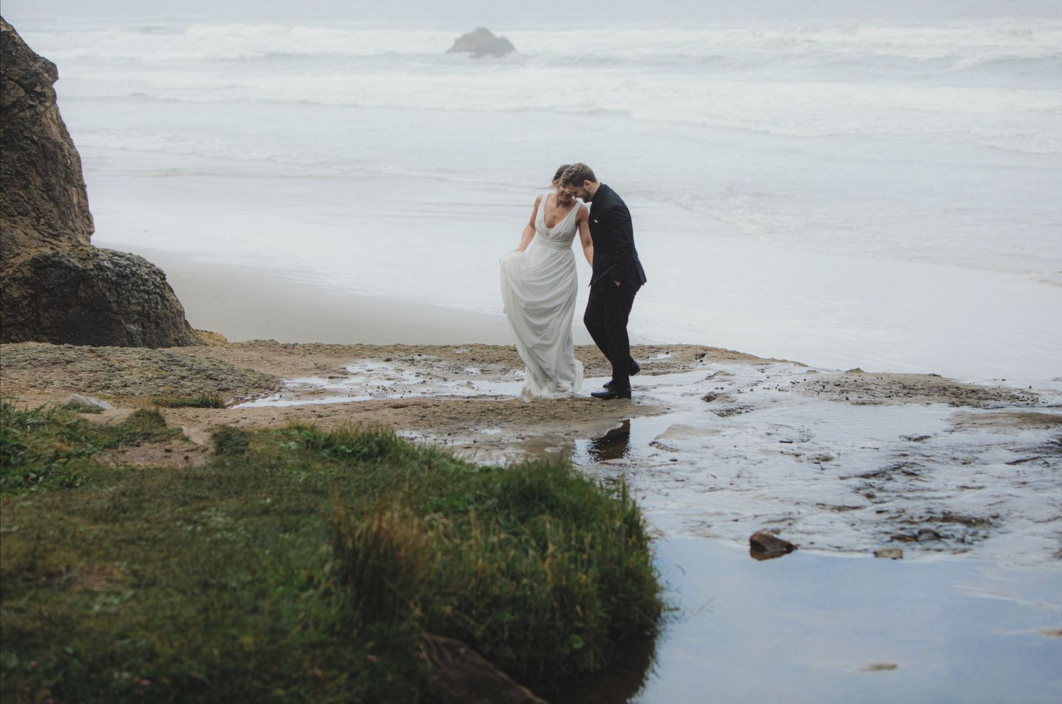 A bride and groom share a romantic kiss on a rocky Pacific coast beach with misty ocean waves in the background.