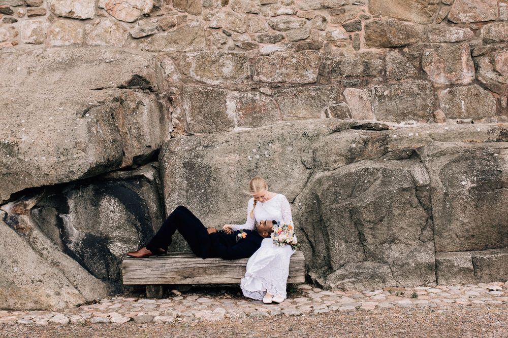 Bride and groom sitting on a wooden bench against a rocky stone wall in a romantic outdoor wedding photo.
