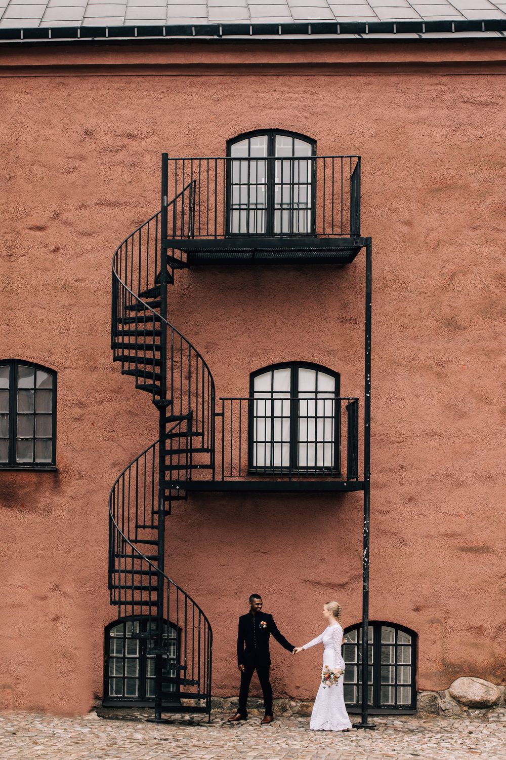 Bride and groom holding hands by a red building with black spiral staircase on their wedding day.