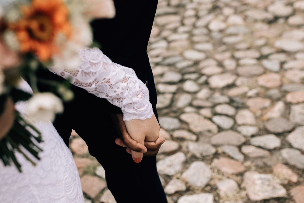 Bride and groom holding hands on cobblestone path, bride wearing lace sleeve holding a bouquet of flowers.