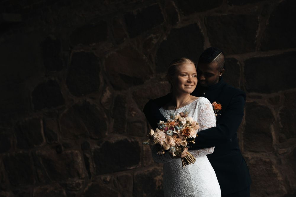 Bride smiles holding a dried floral bouquet as groom embraces her against a dark stone wall.