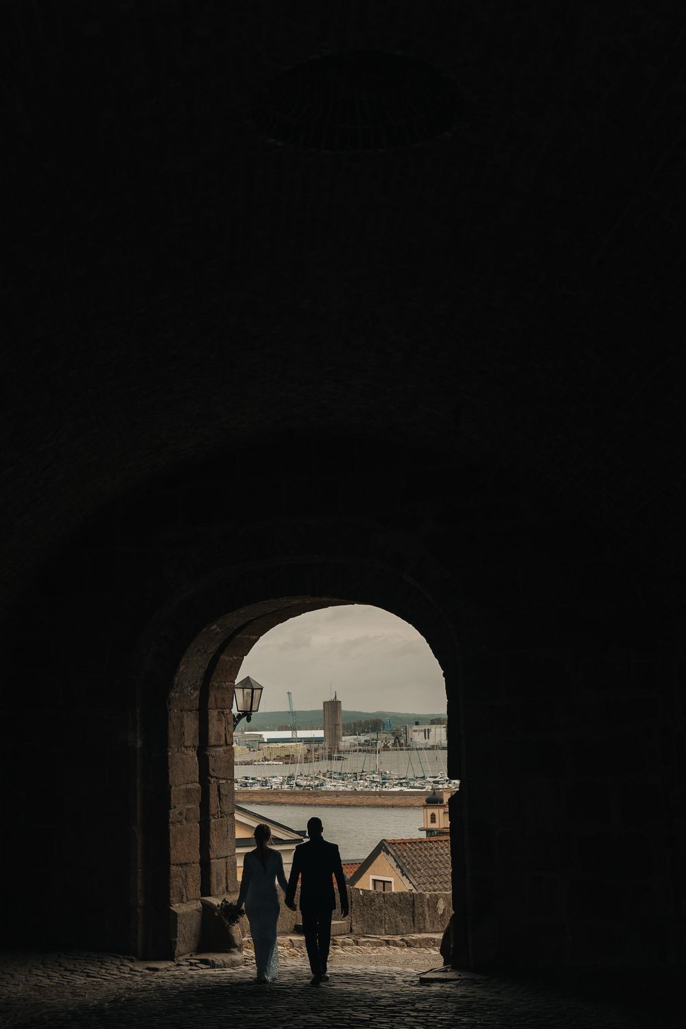 Silhouette of a couple holding hands walking through a dark stone archway with a cityscape view beyond.