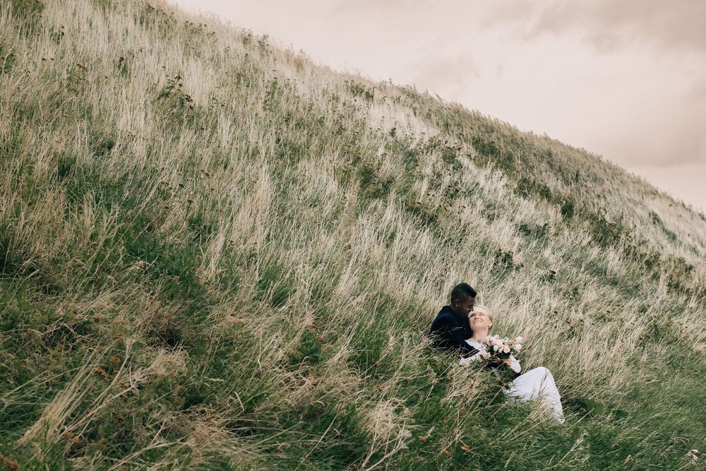 A couple sits together on a lush grassy hillside, the bride holding a bouquet in a serene outdoor wedding setting.