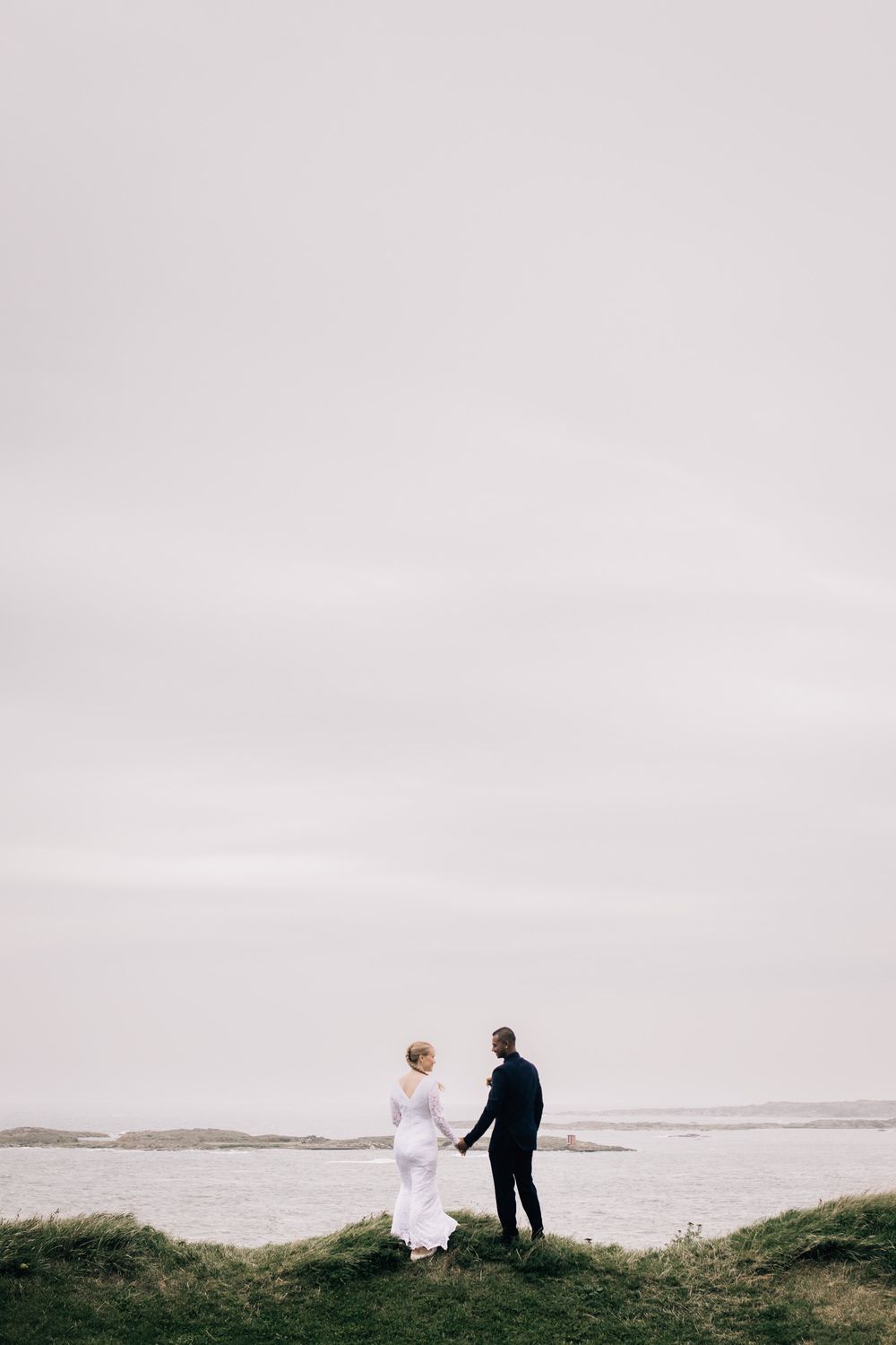 A bride and groom hold hands on a grassy cliff overlooking a misty coastal landscape on their wedding day.