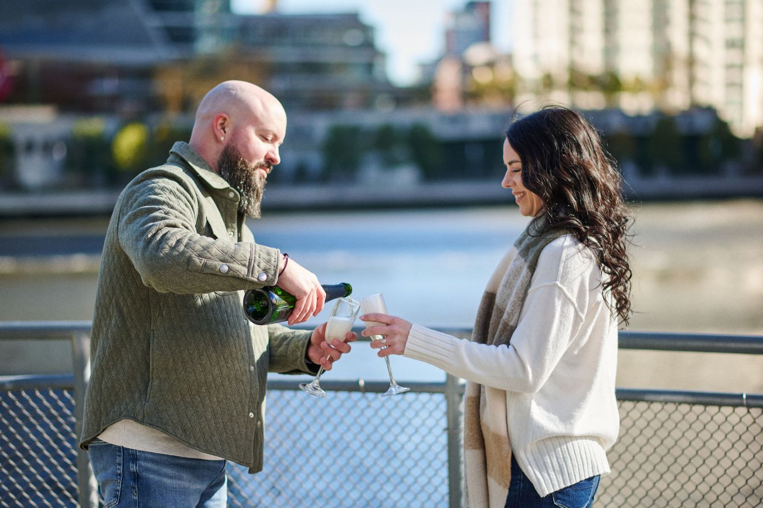 Couple sharing champagne on a waterfront bridge, man pouring wine into woman's glass outdoors.
