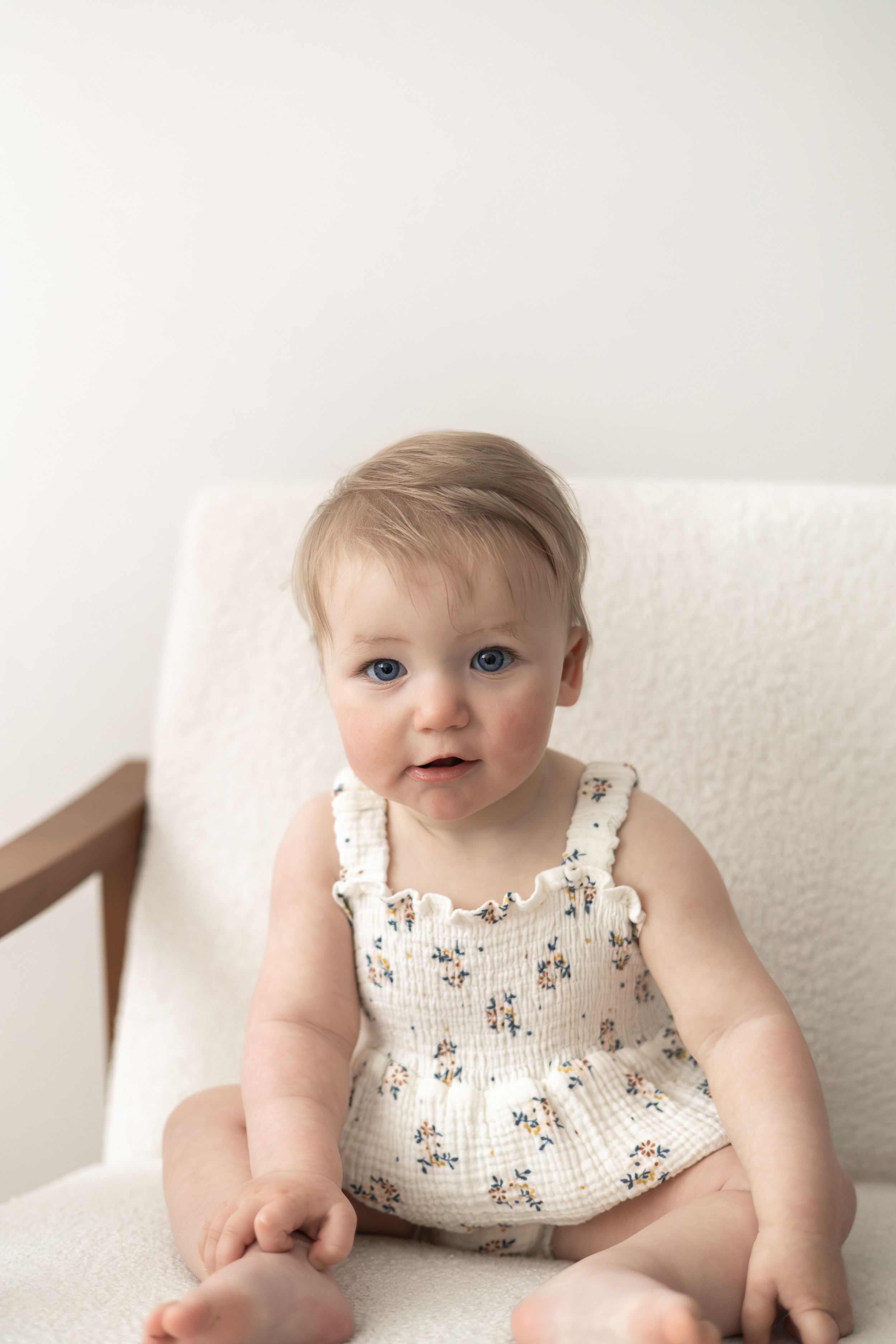 Cute baby with blue eyes sitting in a white chair wearing a floral romper Nottingham natural relaxed baby birthday photoshoot