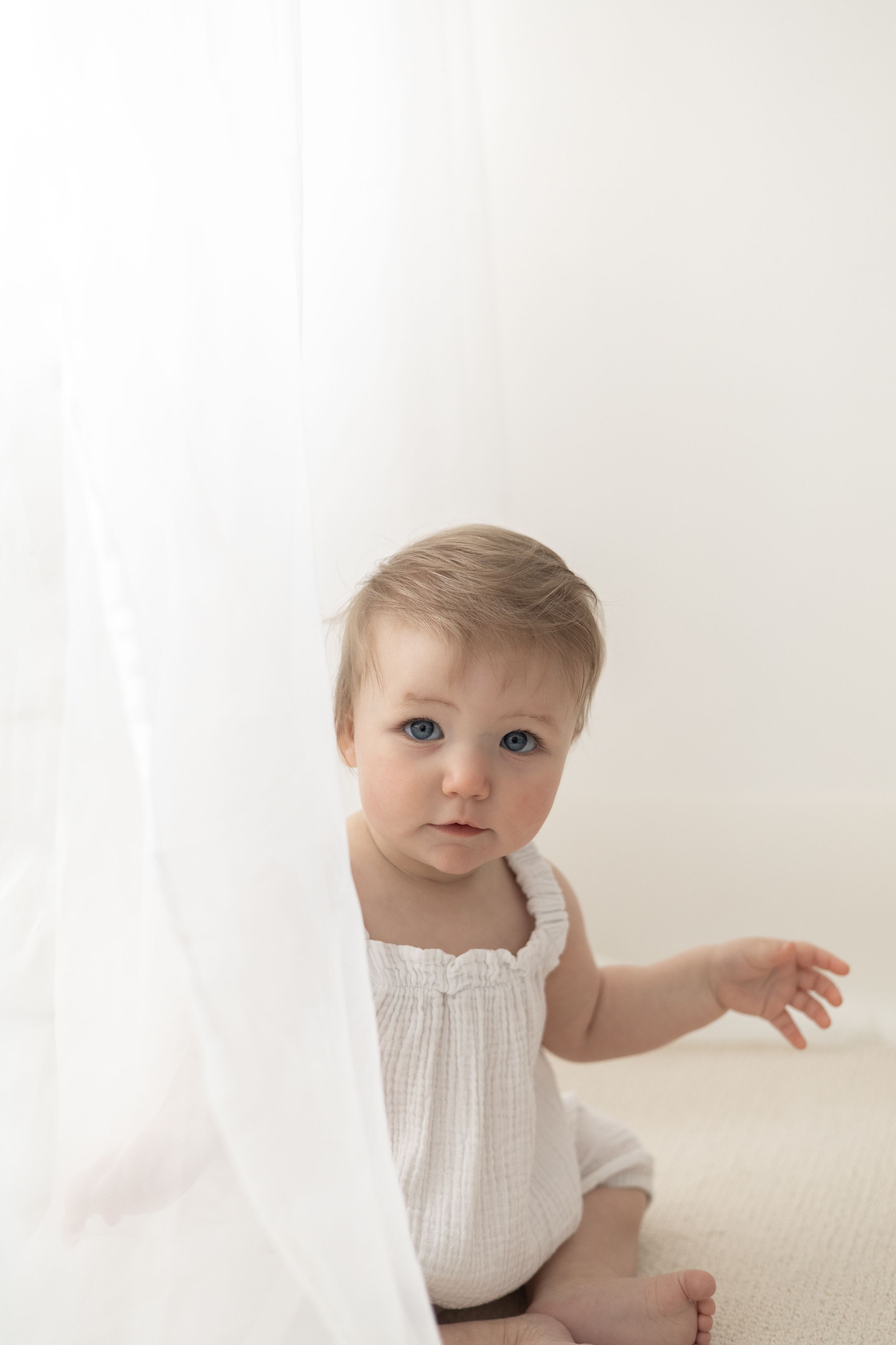 Baby with blue eyes in a white romper peeks around a sheer white curtain Nottingham baby birthday photoshoot natural relaxed