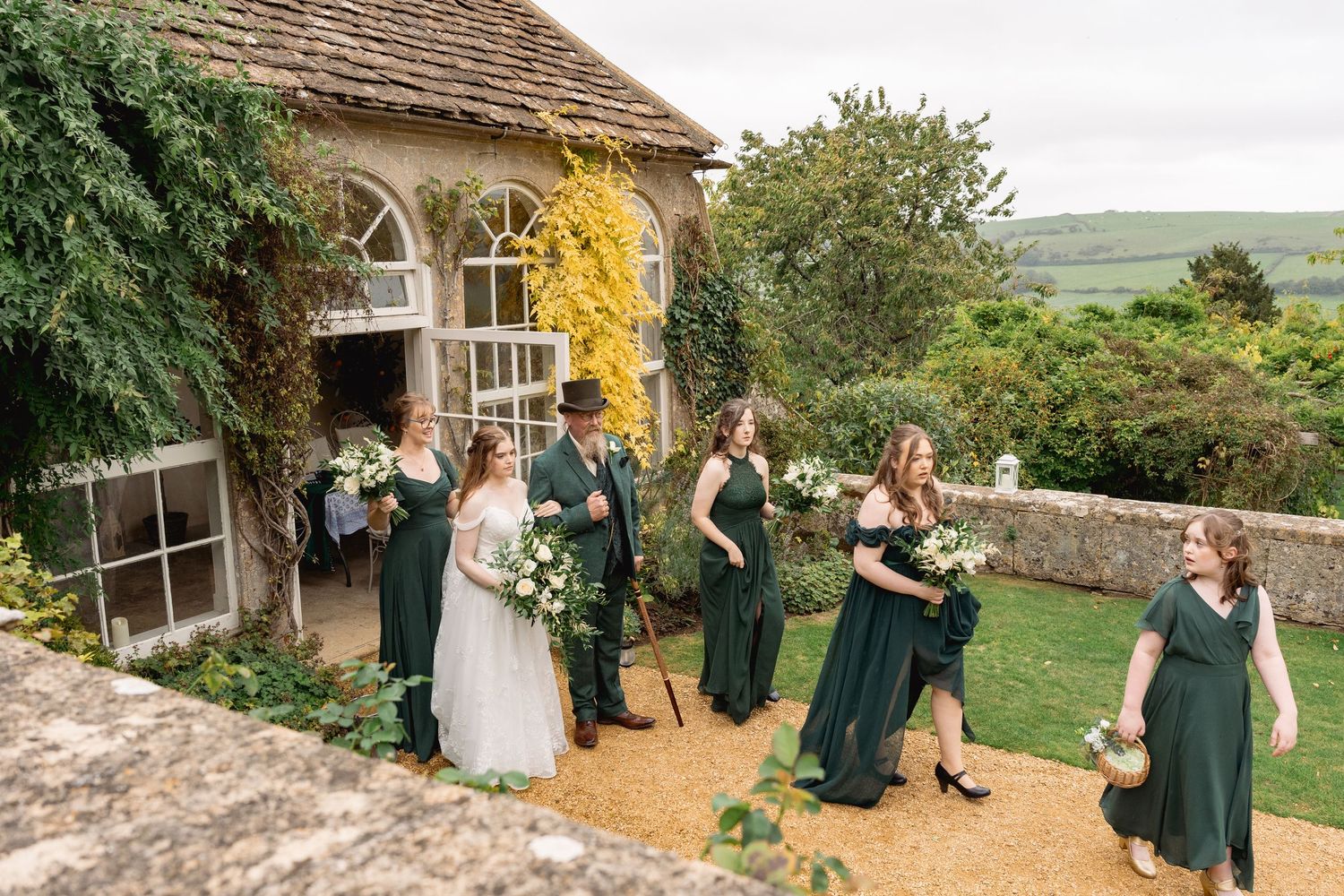 Wedding party in green bridesmaid dresses walking outside a stone venue with scenic countryside views.