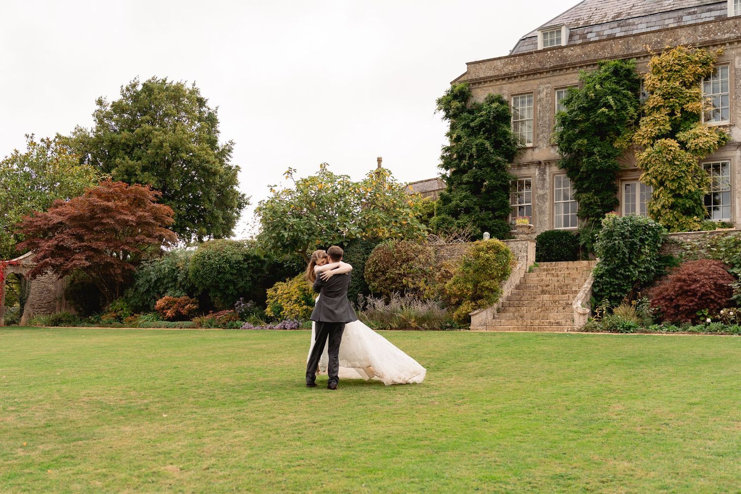 Bride and groom embrace on lush green lawn before grand stone manor house with colorful autumn gardens.