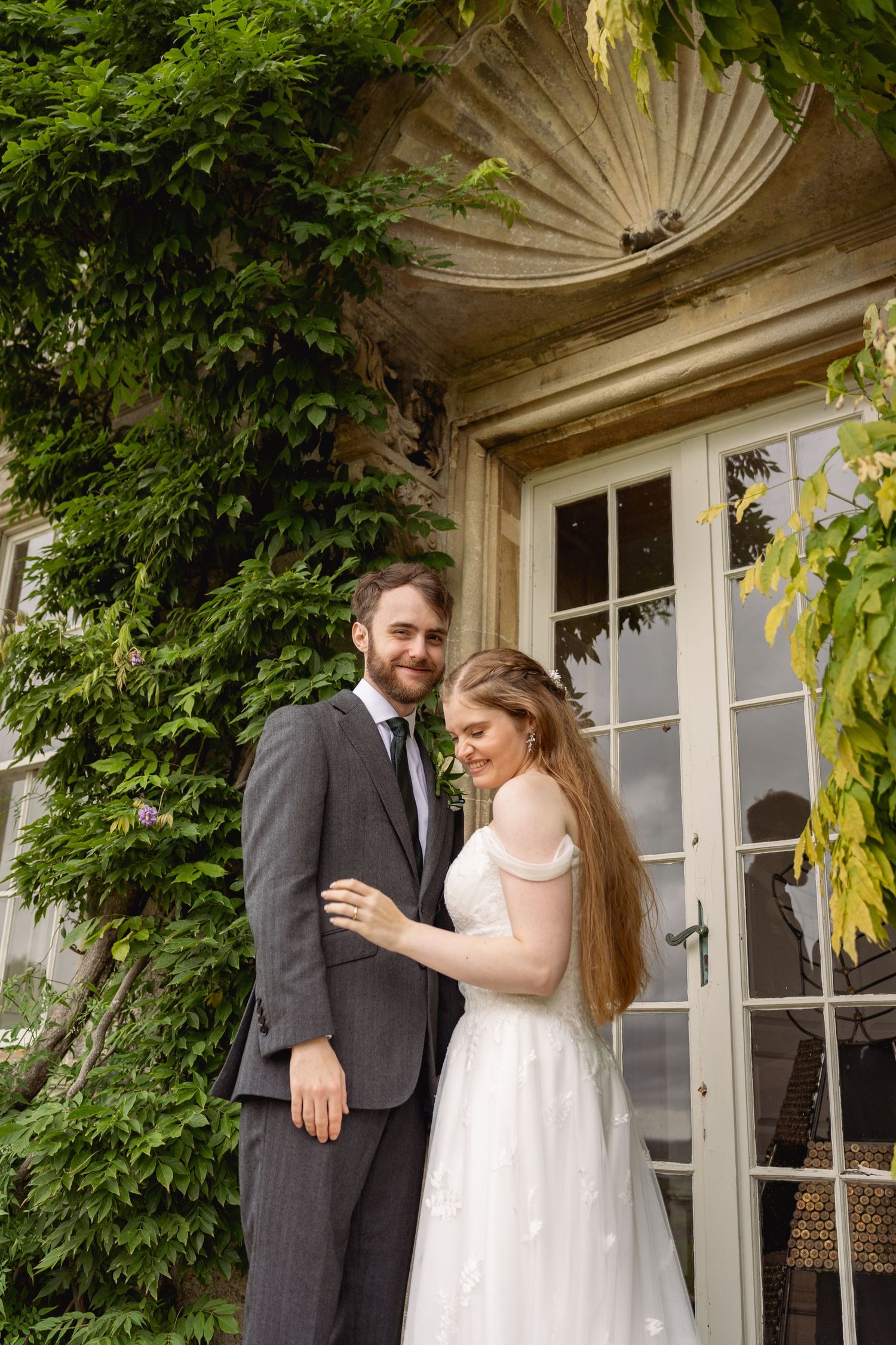 A smiling groom in a gray suit stands with his bride in a white off-shoulder gown outside an ivy-covered stone building.
