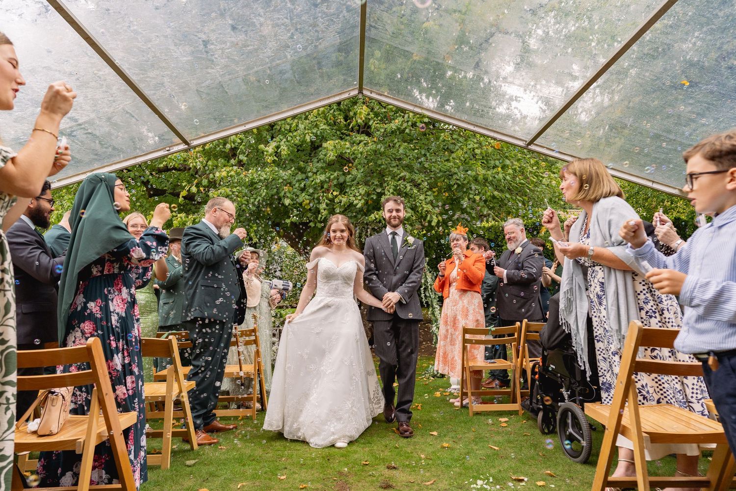 Bride and groom walk down the aisle under a clear marquee tent as guests toss confetti at an outdoor wedding.
