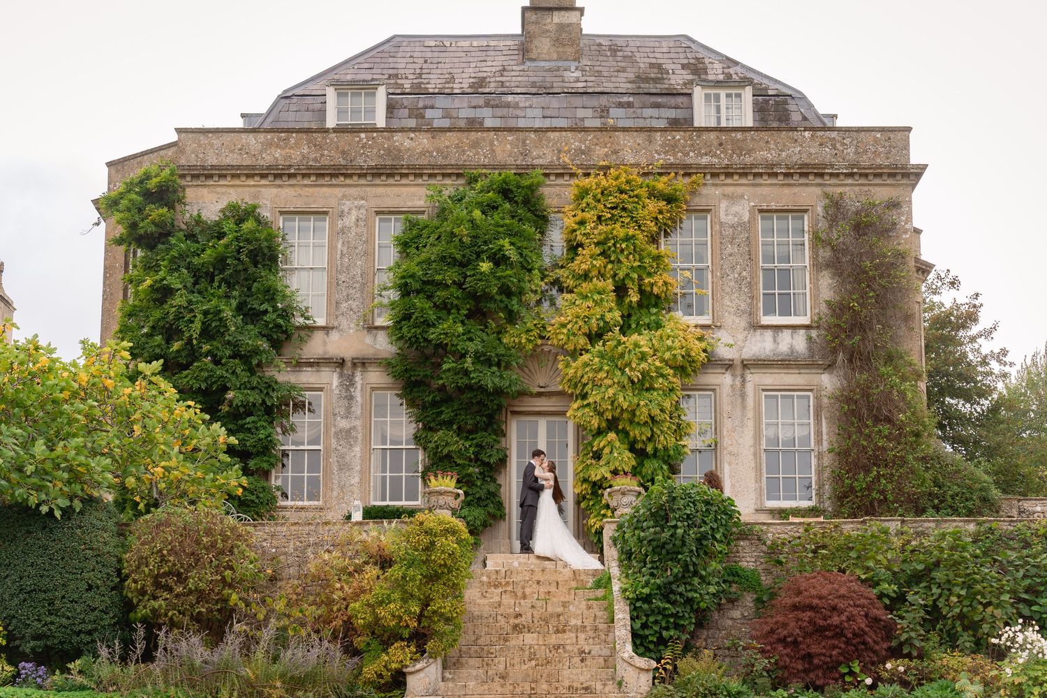 Bride and groom embrace on stone steps of ivy-covered Georgian manor house wedding venue surrounded by lush gardens.