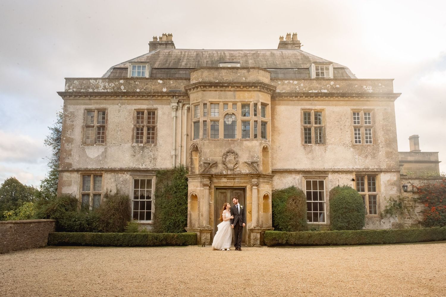 Wedding couple standing before a grand historic English manor house with manicured hedges under a cloudy sky.