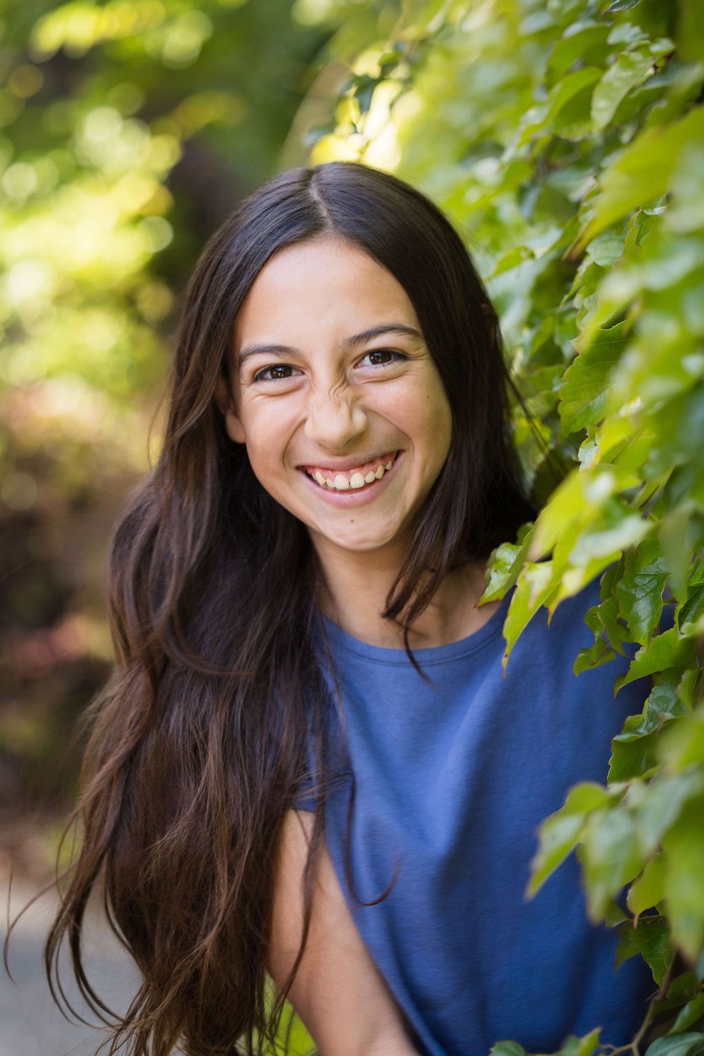 Young woman with long dark hair smiling outdoors among green leaves, wearing a blue top in natural sunlight.