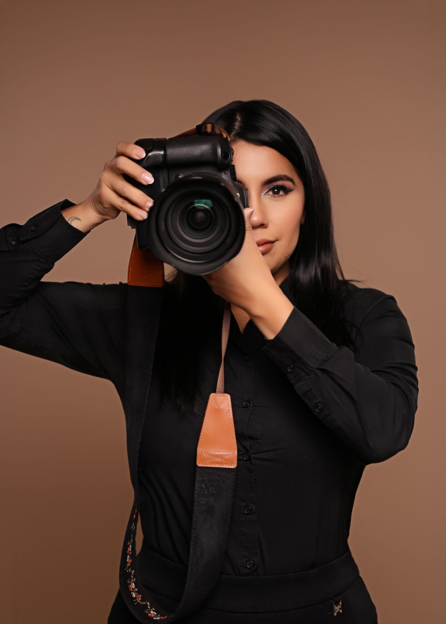 A young woman in black clothing holds a DSLR camera up to her eye against a warm brown background.